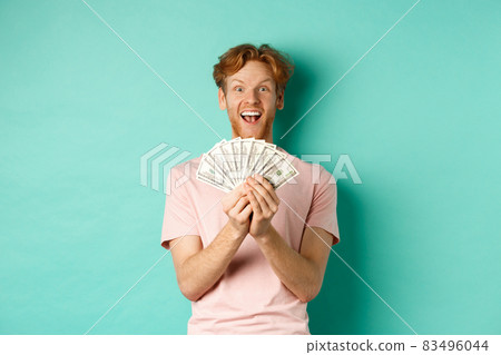 Excited young man winning prize money, counting cash and looking amazed at dollars, standing over turquoise background Excited young man winning prize money, counting cash and looking amazed at dollars, standing over turquoise background 83496044