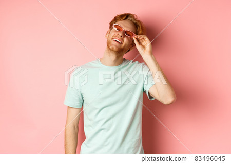 Tourism and vacation concept. Happy young man with red hair relaxing, wearing sunglasses and smiling carefree, standing over pink background Tourism and vacation concept. Happy young man with red hair relaxing, wearing sunglasses and smiling carefree, standing over pink background 83496045