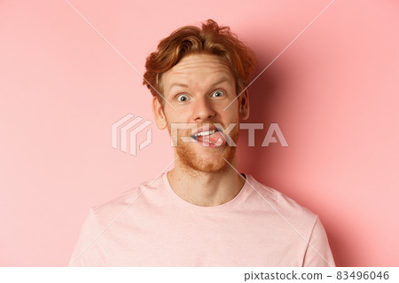 Headshot of funny redhead guy showing tongue, making silly faces at camera, standing joyful against pink background 83496046