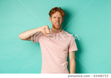 Disappointed redhead man showing thumbs-down and tongue, epxress dislike and disapproval, standing over turquoise background Disappointed redhead man showing thumbs-down and tongue, epxress dislike and disapproval, standing over turquoise background 83496733