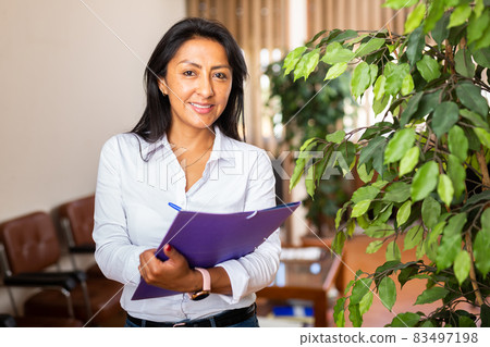 Charming hispanic female office worker standing with documents Charming hispanic female office worker standing with documents 83497198