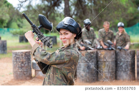 Smiling woman wearing uniform and holding gun ready for playing paintball with friends 83503703