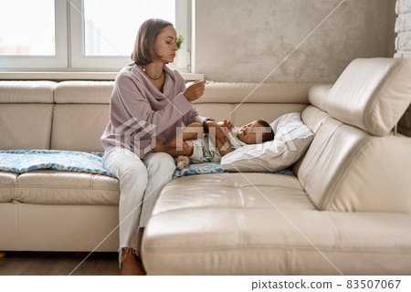 Worried young mother sitting on sofa beside her sick son with high fever. Mom measures temperature 83507067