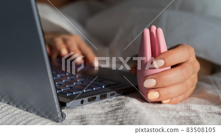 A woman lies in bed holding a clitoral vibrator and watching porn on a laptop. The girl has sex online 83508105