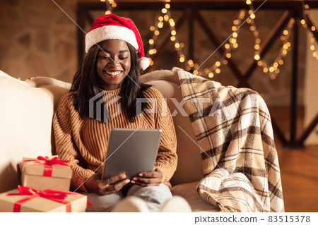 Online Christmas celebration. Black woman in Santa hat making video call, using tablet, shopping for Xmas presents 83515378