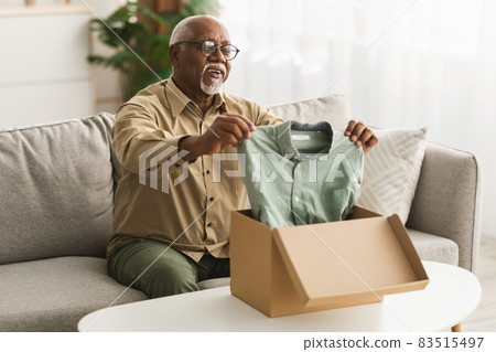 Senior Black Man Unpacking Box Looking At Shirt At Home Senior Black Man Unpacking Box Looking At Shirt At Home 83515497