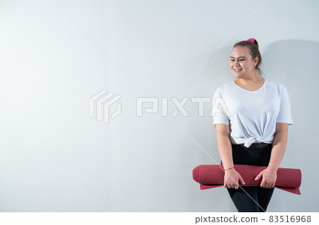 Young fat caucasian woman holding a sport mat. Charming plus size model in sportswear stands on a white background Young fat caucasian woman holding a sport mat. Charming plus size model in sportswear stands on a white background 83516968