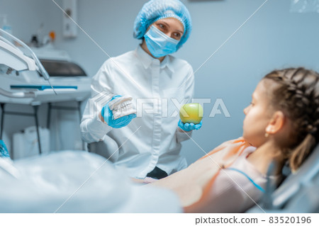 Friendly female dentist orthodontist educating little girl patient using teeth model and apple . 83520196