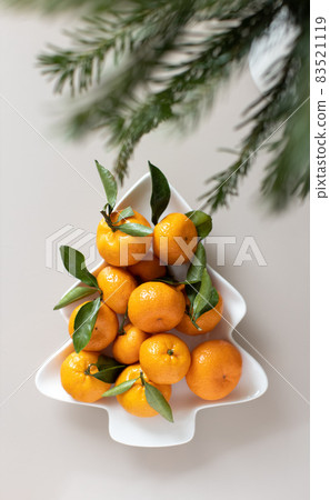 Small ripe tangerines with leaves, on plate in shape of Christmas tree on beige background. Concept winter vitamin food. Moody still life festive composition. For greeting card, book, stories 83521119