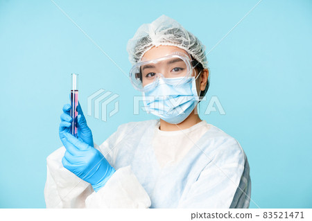 Asian nurse or lab worker in personal protective equipment, showing test sample tube, standing in medical face mask over blue background 83521471