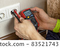 An unrecognizable male technician measures the voltage at the outlet with a digital multimeter. There are only hands in the frame, close-up. Renovation concept, electrician in the apartment 83521743