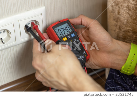 An unrecognizable male technician measures the voltage at the outlet with a digital multimeter. There are only hands in the frame, close-up. Renovation concept, electrician in the apartment 83521743