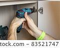 An unrecognizable man installs a door on a wardrobe, fixes furniture with a screwdriver. Only hands are visible. Close-up. Renovation concept, furniture assembly, service sector. Side view 83521747