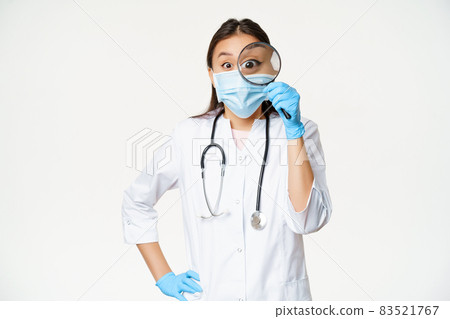 Excited asian medical worker, woman physician looks through magnifying glass surprised, wearing medical face mask and uniform, white background 83521767