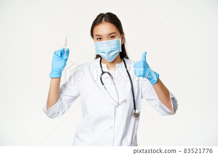 Covid-19 vaccination and healthcare. Asian woman doctor in medical face mask and gloves, holding vaccine syringe and show thumb up, white background 83521776