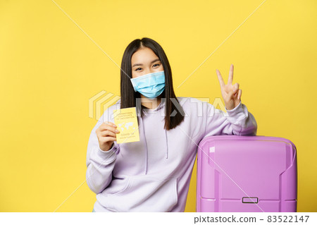 Smiling asian girl tourist in face mask, standing with suitcase, showing covid international vaccination certificate for travellers and peace sign, yellow background Smiling asian girl tourist in face mask, standing with suitcase, showing covid international vaccination certificate for travellers and peace sign, yellow background 83522147