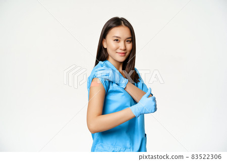 Healthcare workers and vaccination. Young asian nurse in uniform showing arm after vaccinating, thumbs up, standing over white background Healthcare workers and vaccination. Young asian nurse in uniform showing arm after vaccinating, thumbs up, standing over white background 83522306