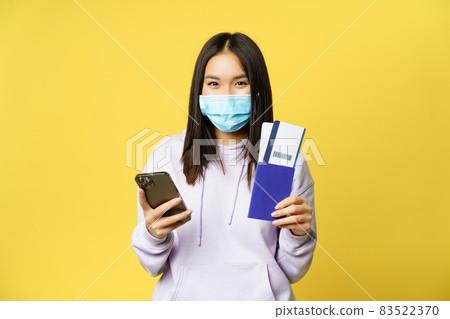 Asian tourist, girl in face mask, holding mobile phone, showing passport and flight tickets, booking hotel for vacation, going abroad, yellow background Asian tourist, girl in face mask, holding mobile phone, showing passport and flight tickets, booking hotel for vacation, going abroad, yellow background 83522370