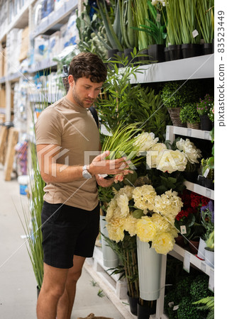 Male customer in casual clothes standing near shelves with potted grass and inspecting grass while shopping in mall Male customer in casual clothes standing near shelves with potted grass and inspecting grass while shopping in mall 83523449