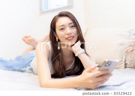 A woman who smiles here while holding a smartphone in bed A woman who smiles here while holding a smartphone in bed 83529422