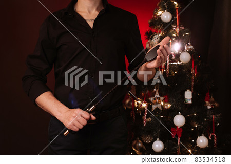 man holds a leather BDSM whip for domination and submission against the background of a Christmas tree man holds a leather BDSM whip for domination and submission against the background of a Christmas tree 83534518