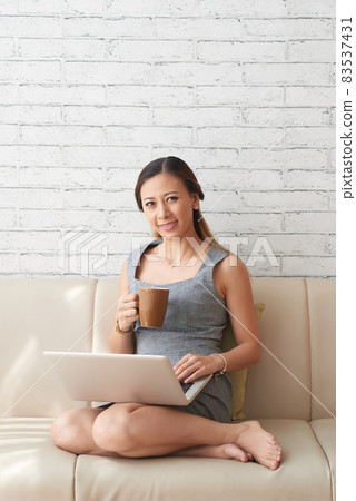 Portrait of smiling young Asian businesswoman in dress sitting with laptop on sofa and drinking coffee while working at home 83537431