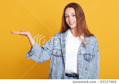 Young girl looking at camera and and spreading hand aside, showing something. Photo of attractive girl shows open palms with something isolated over yellow background. Copy space for advertisment. Young girl looking at camera and and spreading hand aside, showing something. Photo of attractive girl shows open palms with something isolated over yellow background. Copy space for advertisment. 83537469