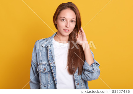 Horizontal shot of young charming woman in denim jacket and white casual shirt standing over yellow background, touches her beautiful long hair, looking directly at camera, being in good mood. 83537486