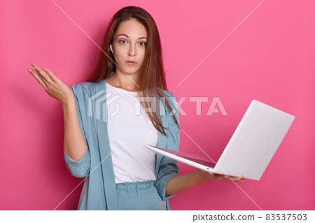 Closeup portrait of astonished and helpless woman posing with hand aside and holding white lap top in hand, lady looking directly at camera, posing isolated over pink studio background. Freelancer. 83537503