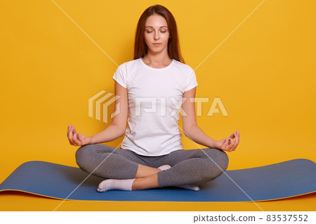 Studio shot of young woman wearing white t shirt and gray leggins, sitting on floor in lotus pose, girl holds hands in praying gesture and keeps eyes closed, posing isolated over yelow background. Studio shot of young woman wearing white t shirt and gray leggins, sitting on floor in lotus pose, girl holds hands in praying gesture and keeps eyes closed, posing isolated over yelow background. 83537552
