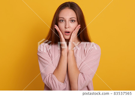 Close up portrait of emotional female being shocked to see something, opens mouth with surprisment and keepspalms near cheek, poses against yellow studio background. People emotions concept. Close up portrait of emotional female being shocked to see something, opens mouth with surprisment and keepspalms near cheek, poses against yellow studio background. People emotions concept. 83537565