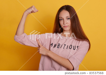 Indoor shot of serious woman with long beautiful hair, wearing pink shirt with inscription, slim female showing her biceps and pointing on it with index finger, posing isolated over yellow background. Indoor shot of serious woman with long beautiful hair, wearing pink shirt with inscription, slim female showing her biceps and pointing on it with index finger, posing isolated over yellow background. 83537568