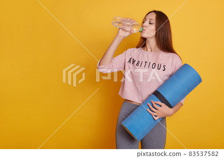 Picture of sporty slim attractive girl with long dark hair, standing and holding yoga mat while drinking water from bottle against yellow wall. Copy space for advertisement or promotional text. Picture of sporty slim attractive girl with long dark hair, standing and holding yoga mat while drinking water from bottle against yellow wall. Copy space for advertisement or promotional text. 83537582