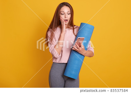 Sporty woman holding smartphone in hand, standing with shocked facial expression and opened mouth. Healthy young female check social network before workout, isolated over yellow studio wall. Sporty woman holding smartphone in hand, standing with shocked facial expression and opened mouth. Healthy young female check social network before workout, isolated over yellow studio wall. 83537584