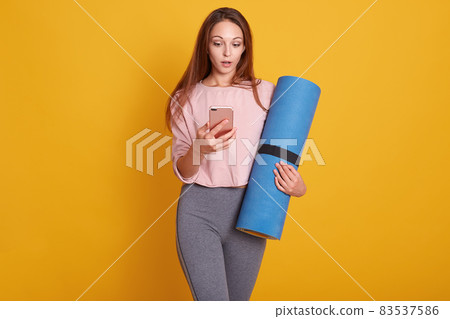 Indoor shot of young yoga instructor stand holding mat, checking mail on smartphone, fit female wearing sportswear use mobile phone, waiting for training in fitness studio, isolated over yellow wall. Indoor shot of young yoga instructor stand holding mat, checking mail on smartphone, fit female wearing sportswear use mobile phone, waiting for training in fitness studio, isolated over yellow wall. 83537586