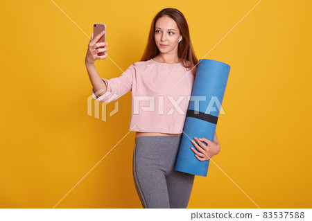 Image of young and active dark haired woman posing with blue yoga mat and makes selfie via phone in studio isolated over yellow background, lady wearig stylish sportwear. Fitness and diet concept. Image of young and active dark haired woman posing with blue yoga mat and makes selfie via phone in studio isolated over yellow background, lady wearig stylish sportwear. Fitness and diet concept. 83537588