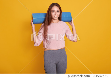 Indoor shot of young happy beautiful Caucasian woman,looks smiling at camera, holding yoga mat over her shoulder, ready for gym, wearing stylish sportwear. Fitness, diet, healthy lifestyle concept. Indoor shot of young happy beautiful Caucasian woman,looks smiling at camera, holding yoga mat over her shoulder, ready for gym, wearing stylish sportwear. Fitness, diet, healthy lifestyle concept. 83537589