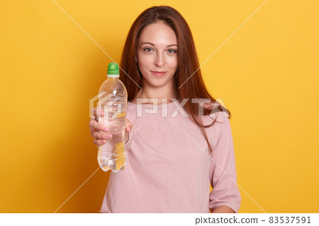 Portrait of beautiful sports woman showing bottle of water isolated on yellow background, girl wearing pink shirt, attractive famale having long brown hair, lady looking smiling directly at camera. Portrait of beautiful sports woman showing bottle of water isolated on yellow background, girl wearing pink shirt, attractive famale having long brown hair, lady looking smiling directly at camera. 83537591