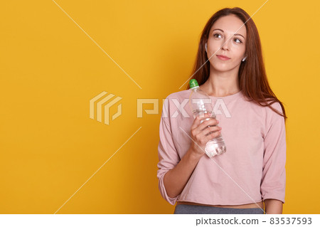 Young athletic woman wearing wireless headphones and rose sportswear holds bottle of water in hands, looking up, ready for running, isolated over yellow wall, copy space for promotion or advertismant. Young athletic woman wearing wireless headphones and rose sportswear holds bottle of water in hands, looking up, ready for running, isolated over yellow wall, copy space for promotion or advertismant. 83537593