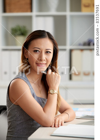 Portrait of smiling confident young female Asian manager in golden wristwatch sitting at desk and holding pen in office 83537691