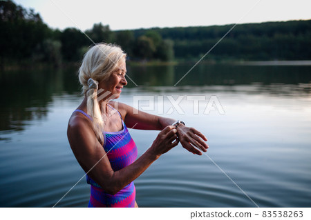 Portrait of active senior woman swimmer standing and setting smartwatch outdoors in lake. 83538263