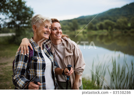 Happy senior mother hiker embracing with adult daughter when looking at lake outdoors in nature Happy senior mother hiker embracing with adult daughter when looking at lake outdoors in nature 83538286