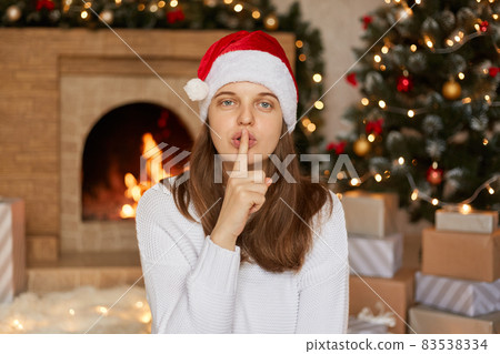 Woman with Santa hat making quiet gesture while looking directly at camera and keeping finger near lips, girl with straight hair posing in living room with x-mas tree and fireplace on background. 83538334
