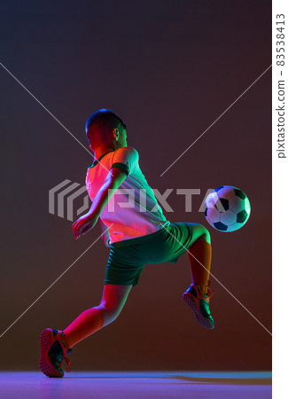 Full-length portrait of boy, child in motion training football isolated over gradient background in neon light. Back view 83538413