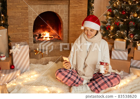 Photo of positive lady sitting with cellphone and holding beverage mug in house indoors with christmas x-mas ornament, wearing santa hat, scarf and checkered pants, having video call. Photo of positive lady sitting with cellphone and holding beverage mug in house indoors with christmas x-mas ornament, wearing santa hat, scarf and checkered pants, having video call. 83538620