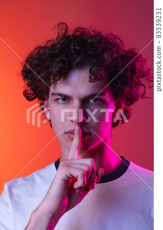 Cropped portrait of young man in white T-shirt with silent expression isolated over gradient pink orange background in neon lights Cropped portrait of young man in white T-shirt with silent expression isolated over gradient pink orange background in neon lights 83539231