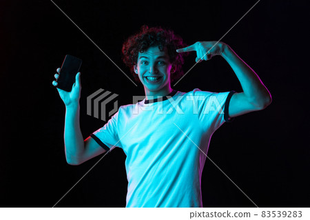Cropped portrait of young man in white T-shirt pointing at phone screen isolated over black background in neon lights. Happy and excited look 83539283