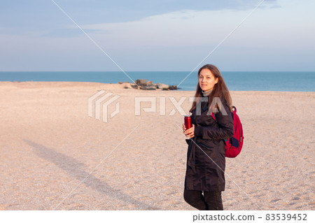 Happy young woman in black raincoat empty autumn sea beach. Smiling girl drinking tea with thermo can bottle. Lifestyle Happy young woman in black raincoat empty autumn sea beach. Smiling girl drinking tea with thermo can bottle. Lifestyle 83539452
