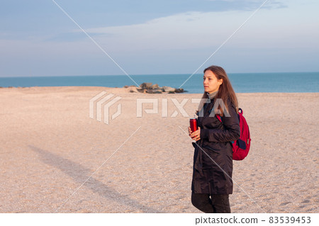 Happy young woman in black raincoat empty autumn sea beach. Smiling girl drinking tea with thermo can bottle. Lifestyle 83539453