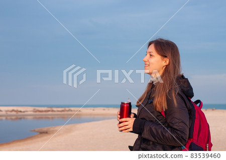 Happy young woman in black raincoat empty autumn sea beach. Smiling girl drinking tea with thermo can bottle. Lifestyle 83539460